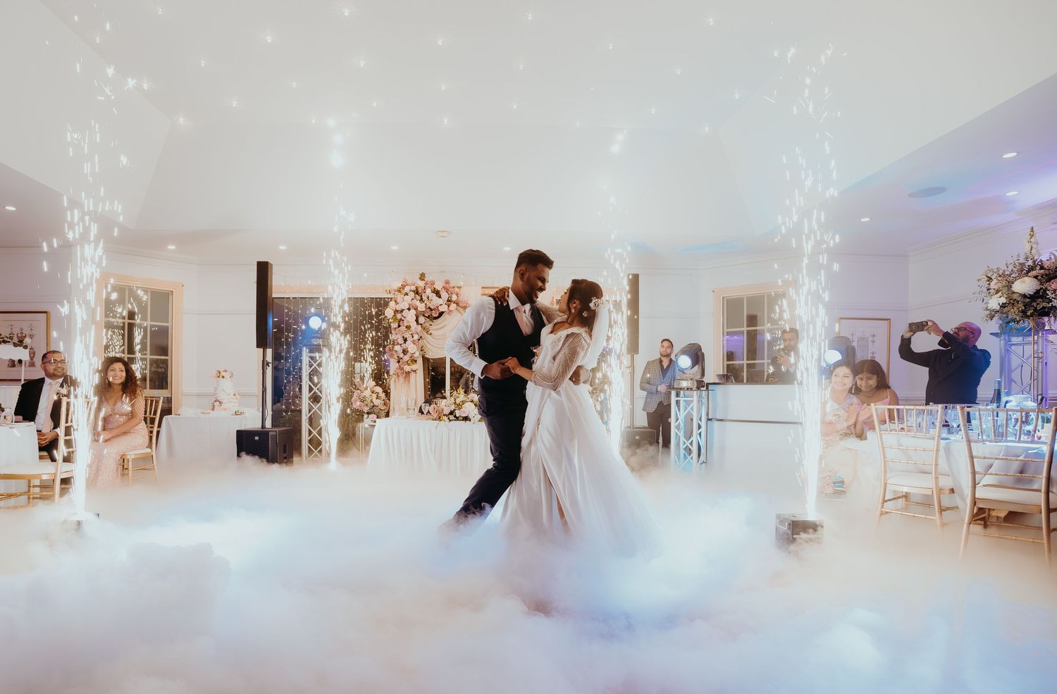 A couple shares their first dance at a wedding reception in an elegant white venue with fog and twinkling lights.