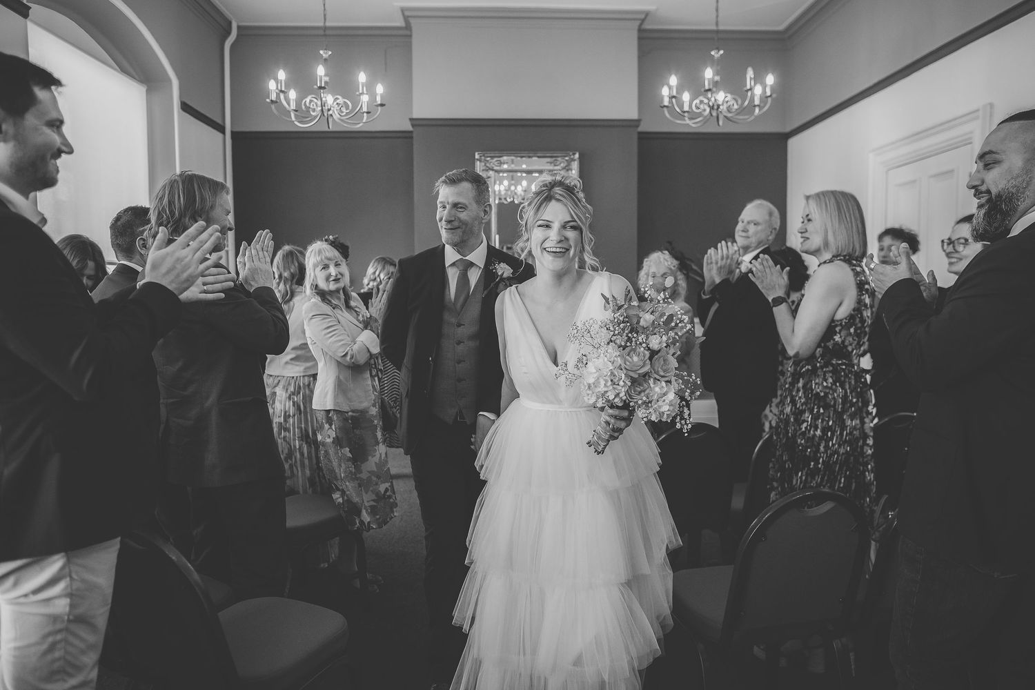 A vintage red double decker bus serves as backdrop for a wedding celebration with guests gathered outside on a sunny day.