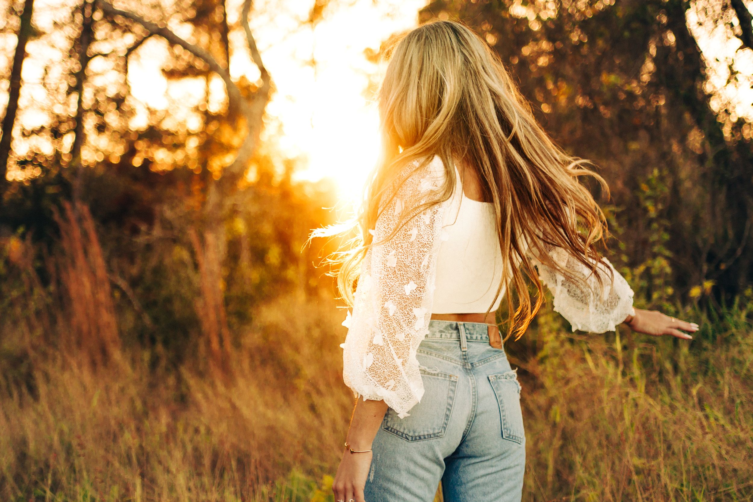Blonde-haired figure in lace top and jeans stands silhouetted against golden sunset in rustic field.