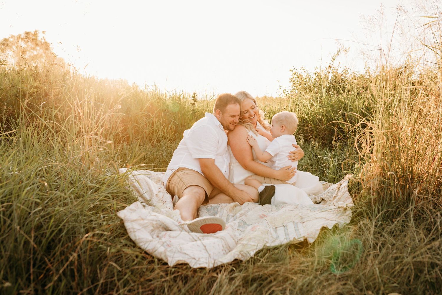 Family relaxing together on white blanket in golden field at sunset.