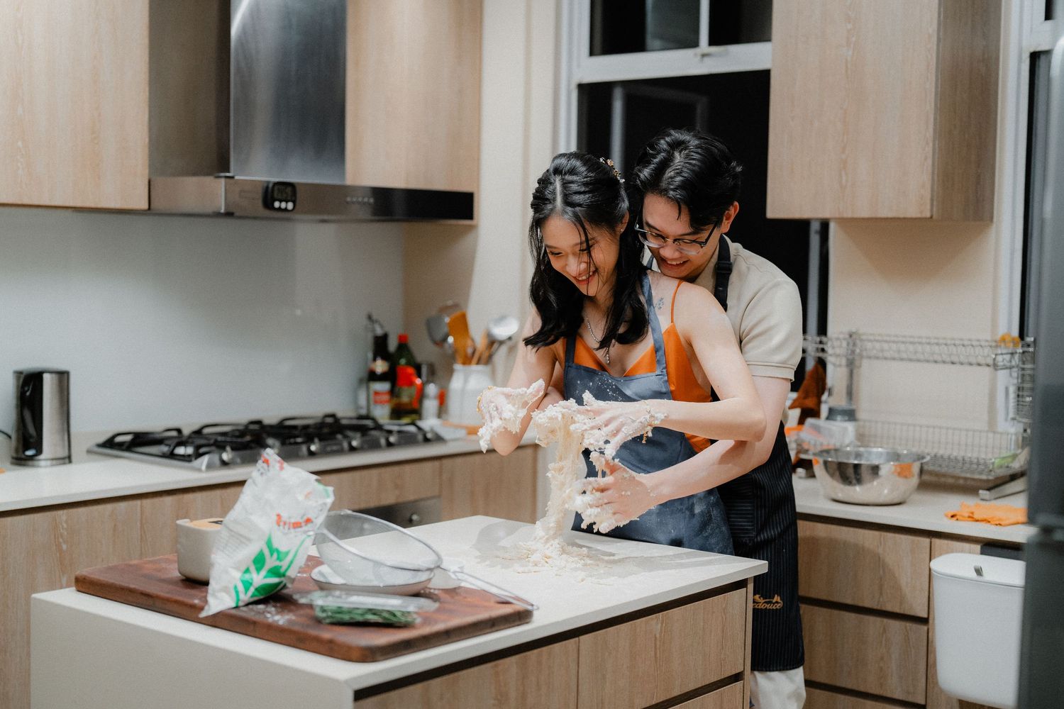 Two people cooking together in a modern kitchen with light wood cabinets and white countertops.