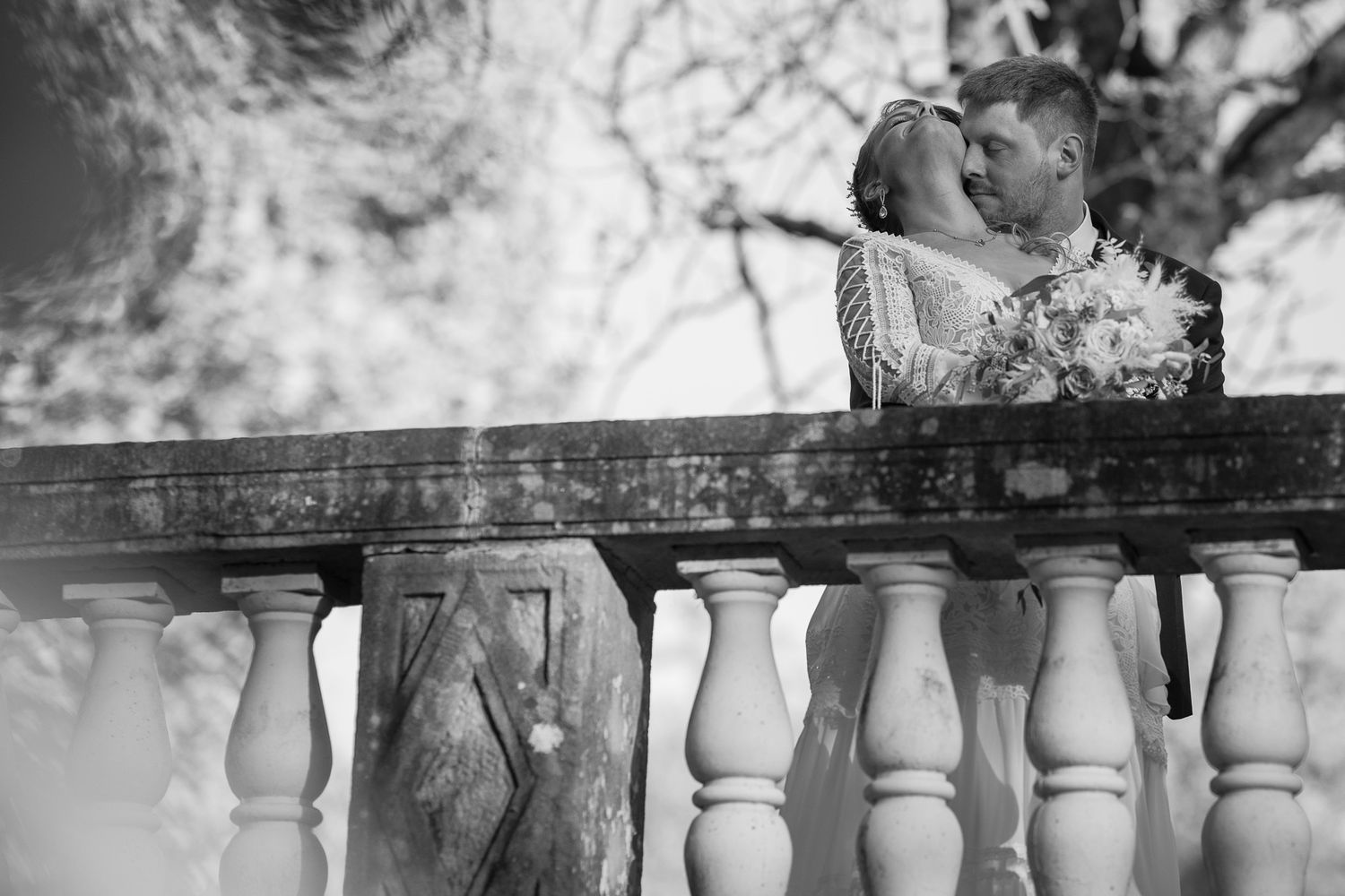 A romantic black and white wedding photo showing a couple embracing on a balcony with decorative stone balusters.