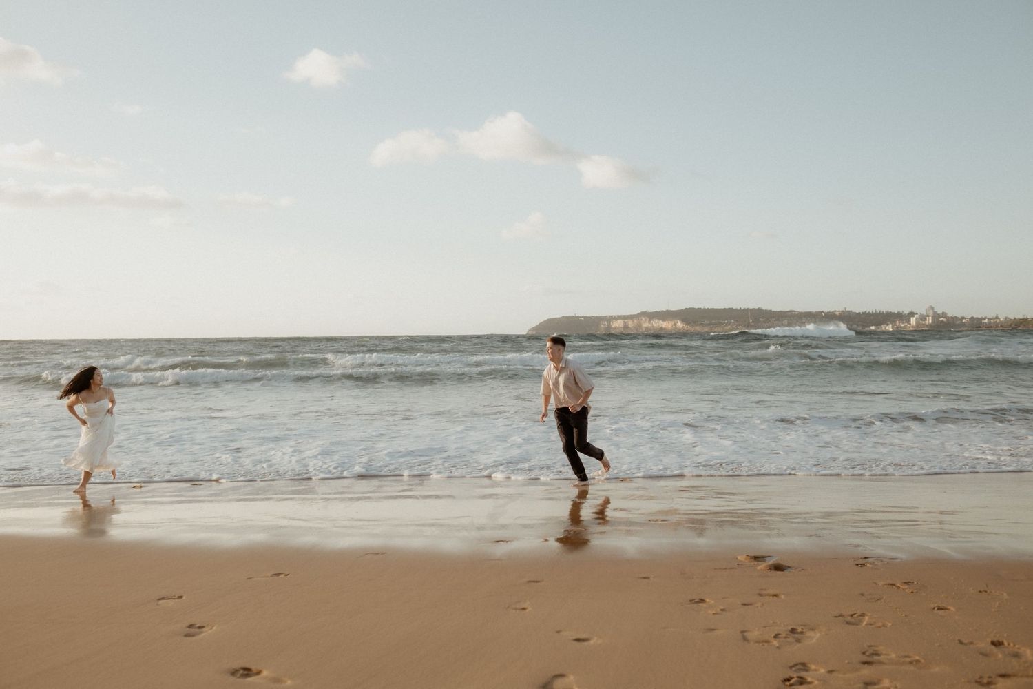 Two people running and playing on a sunny beach with waves in the background.