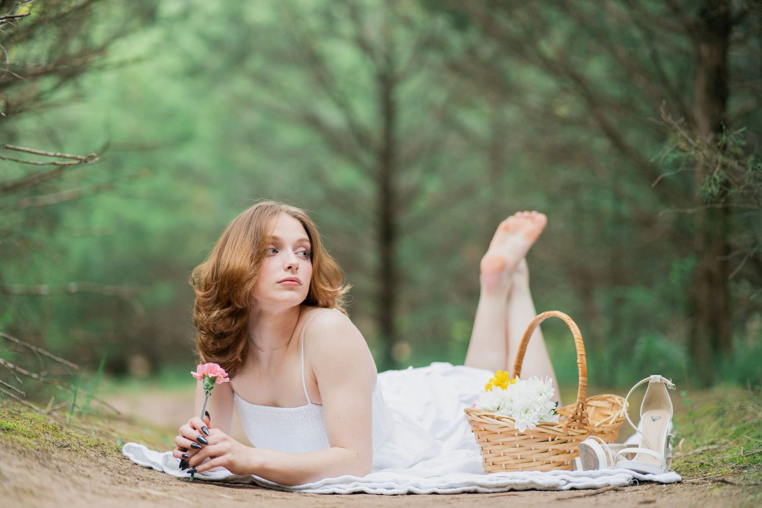 Young woman in white dress lying on blanket in pine forest holding pink flower with picnic basket.