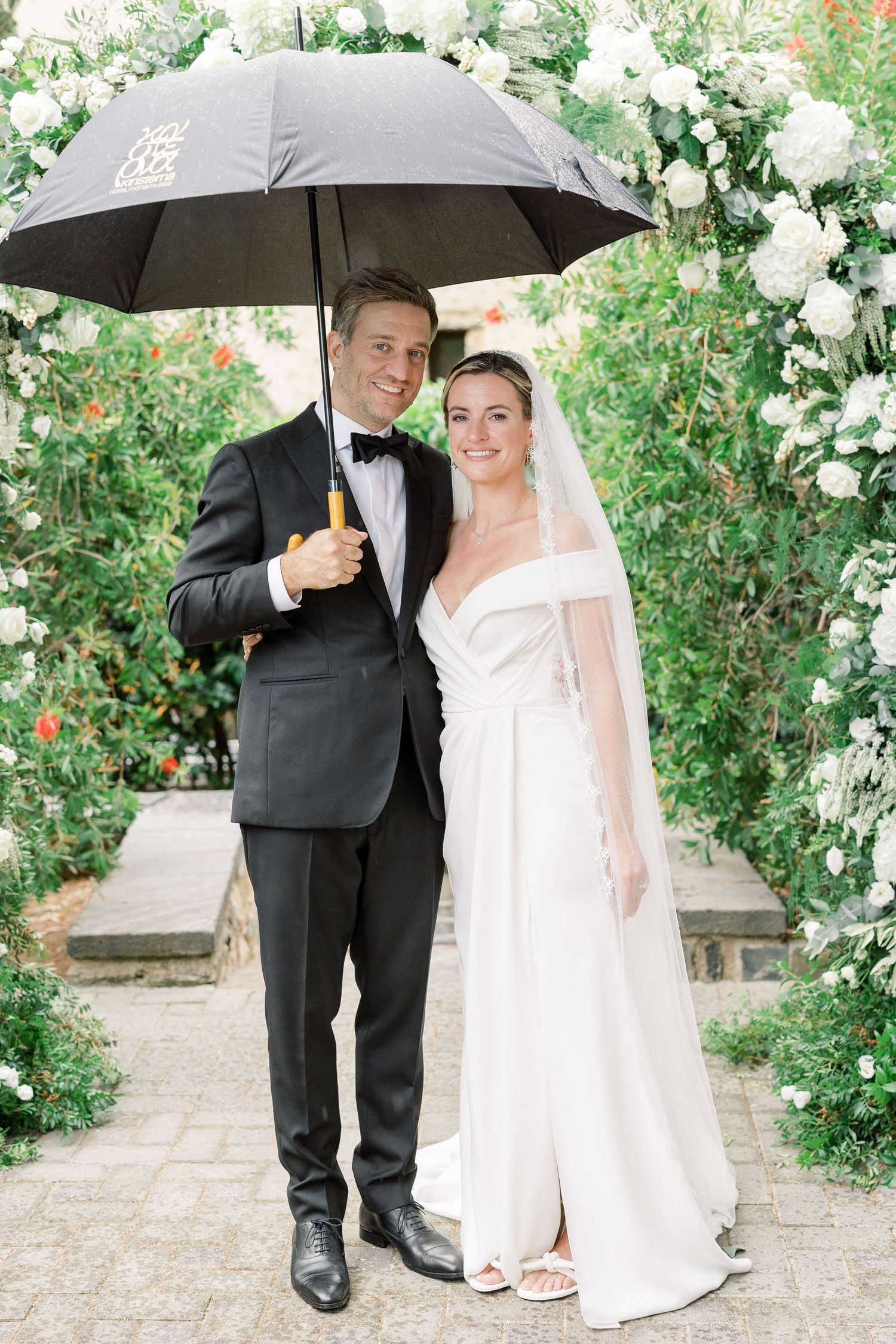 Wedding couple under black umbrella surrounded by white floral archway in garden setting.