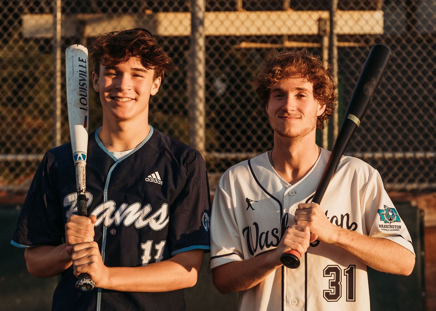 Two baseball players pose with bats wearing contrasting home and away jerseys at a baseball field.