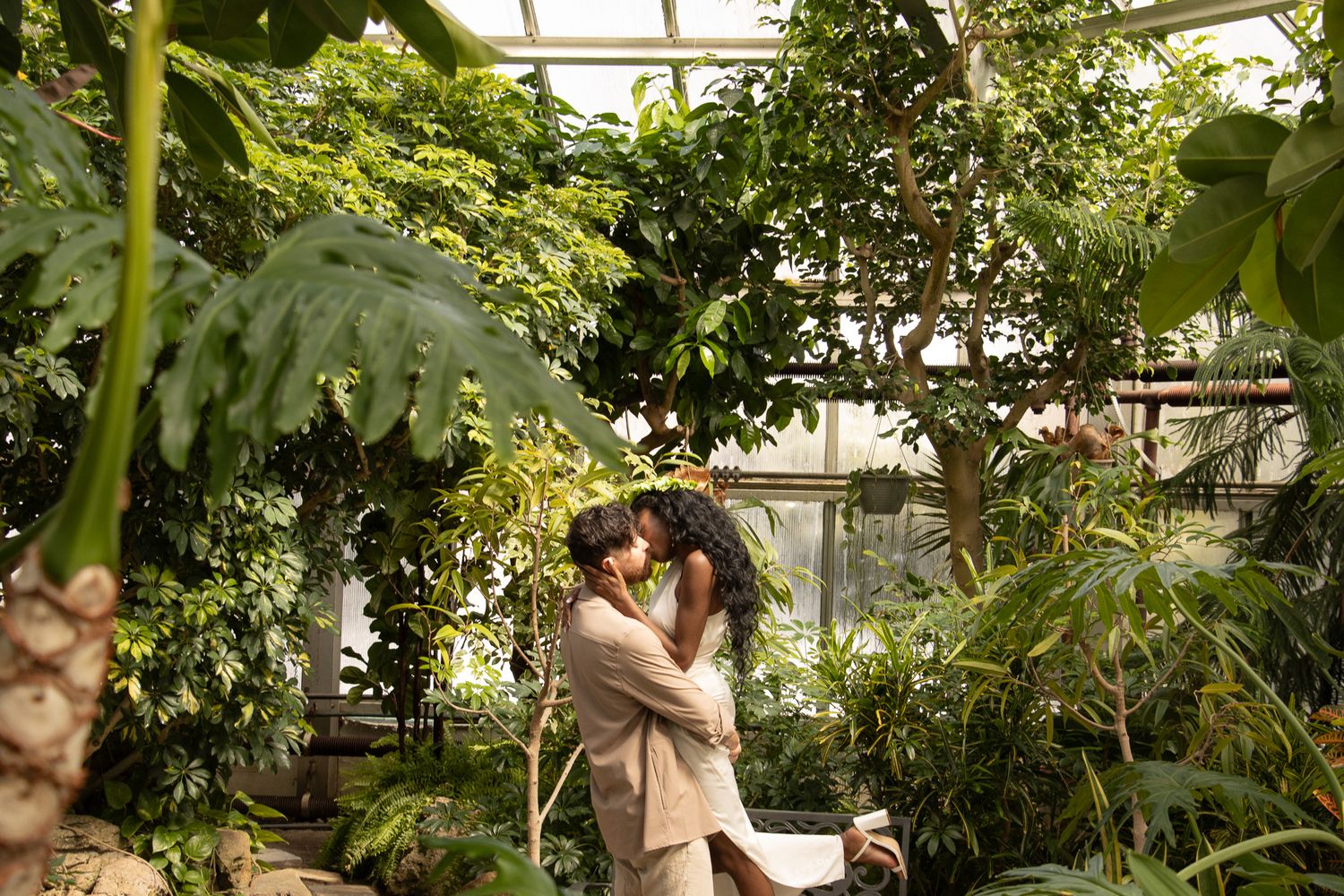 A joyful couple embraces outdoors, surrounded by lush greenery. The woman, wearing a white dress and a leafy crown.