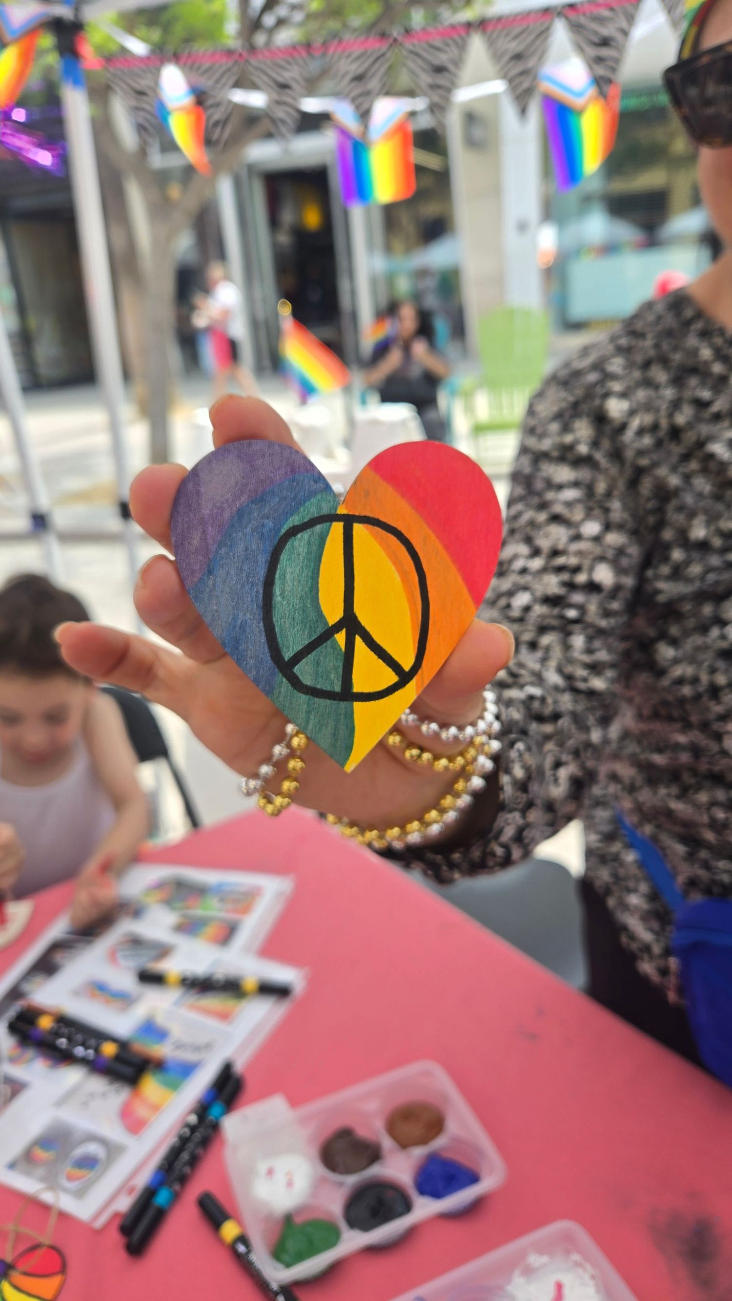 Hand holding a rainbow heart with peace sign at Pride event, with art supplies and rainbow decorations in background.
