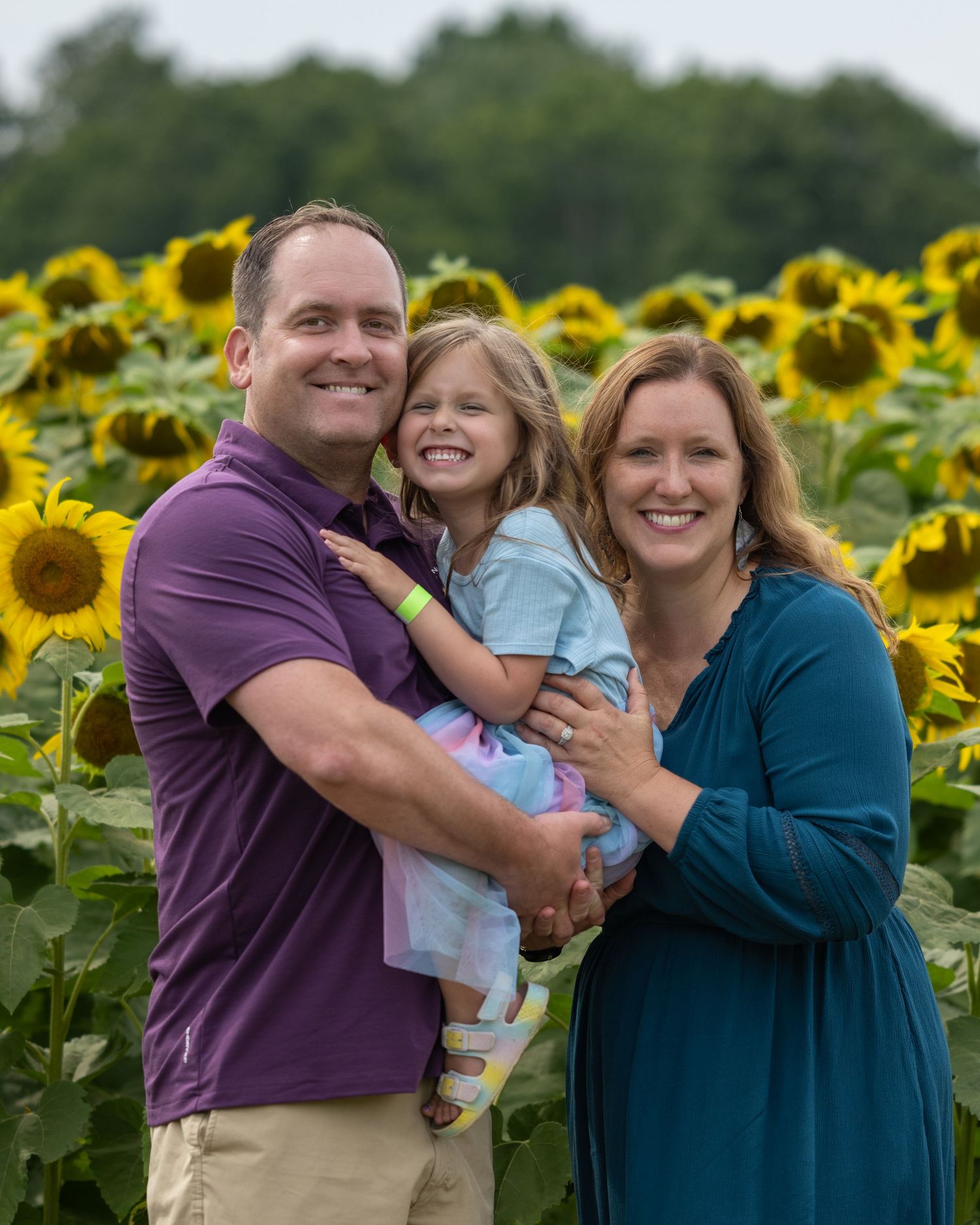 A smiling family of three poses in a vibrant sunflower field, with a man in purple holding a young girl beside a woman in teal.