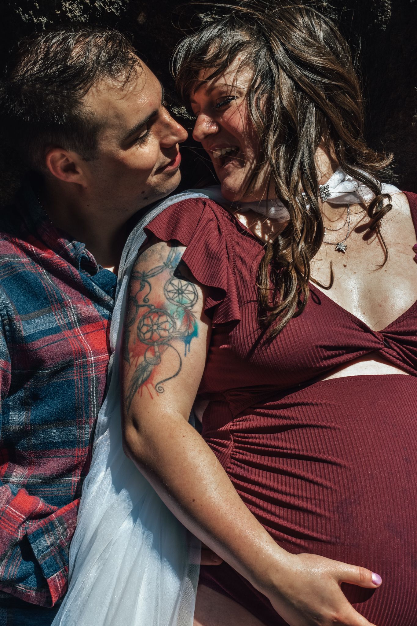 A man with tattoos and a woman in a burgundy dress share an intimate moment, with the woman showing a pregnant belly.