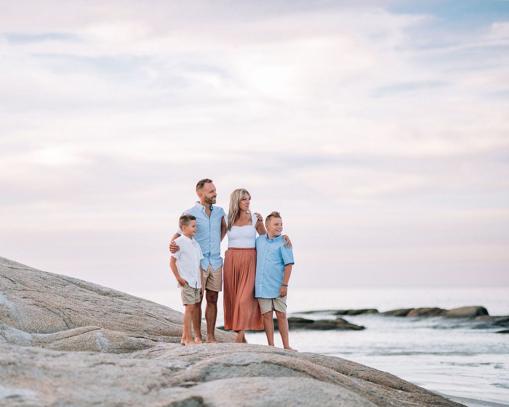 Family of four stands on coastal rocks at sunset, wearing coordinating blue and neutral outfits, with ocean waves behind them.