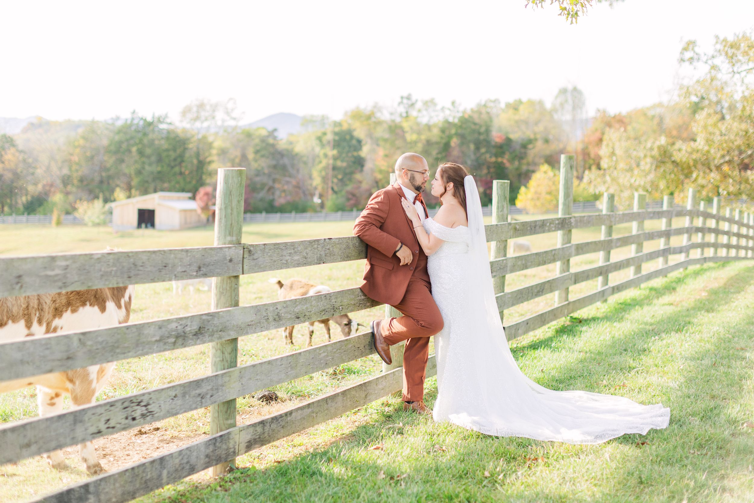 Bride and groom share a romantic kiss leaning against a rustic wooden fence on a sunny fall day at an outdoor farm venue.