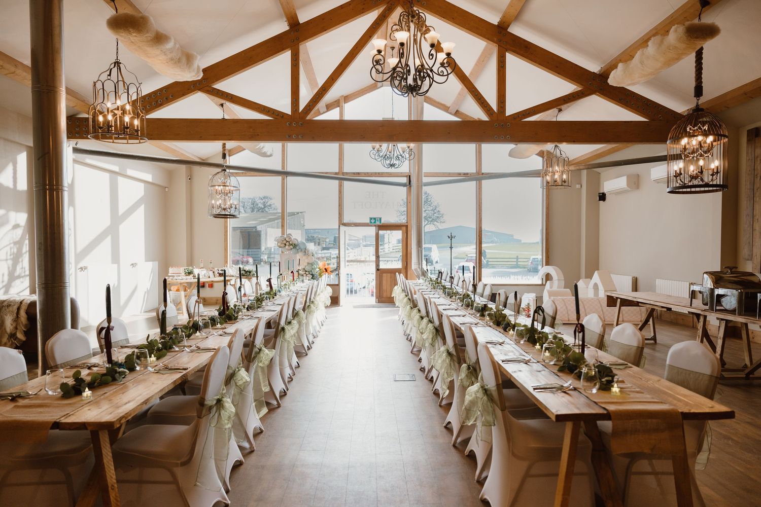 Elegant wedding reception hall with long wooden tables, greenery garlands, chandeliers, and mountain views through large windows.