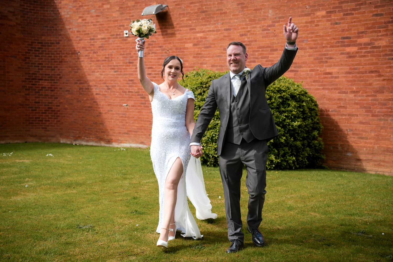 Joyful newlyweds celebrating outdoors, bride in white dress holding bouquet, groom in gray suit, raising hands in triumph.
