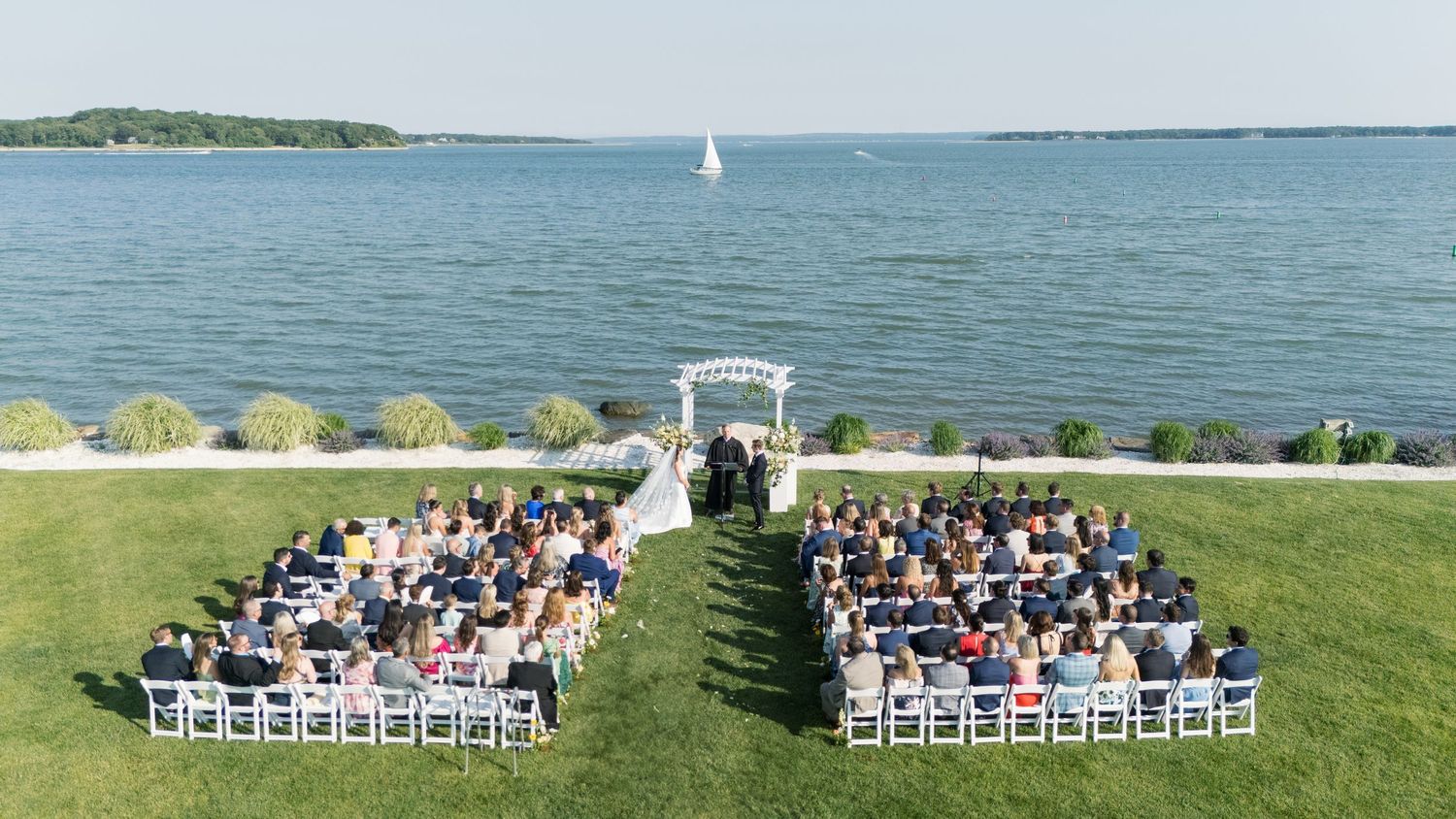 Outdoor waterfront wedding ceremony with white chairs on green lawn, guests seated facing floral arch with sailboat in background.