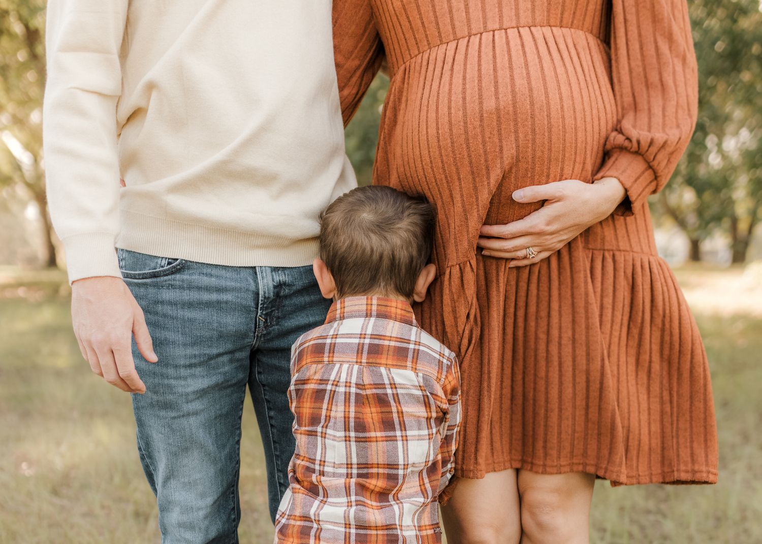Pregnant woman in rust dress with partner and young boy in plaid shirt standing outdoors during maternity photo session.