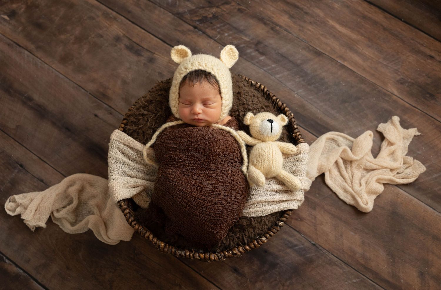 Newborn baby sleeping in a wicker basket wearing a bear bonnet, wrapped in brown knit, with a teddy bear on wood floor.