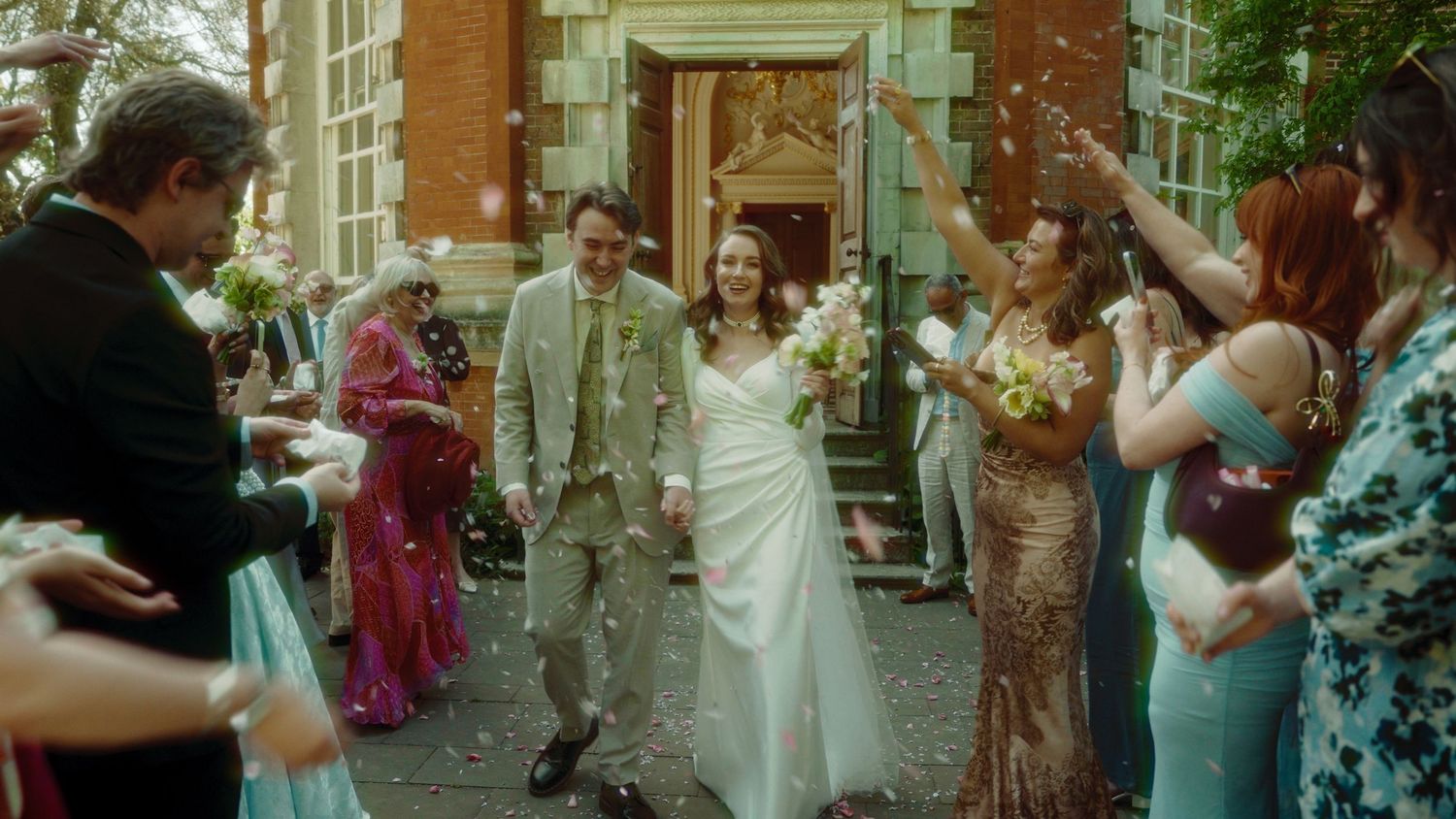 Joyful newlyweds walk through confetti shower outside brick venue, surrounded by smiling guests in elegant attire.