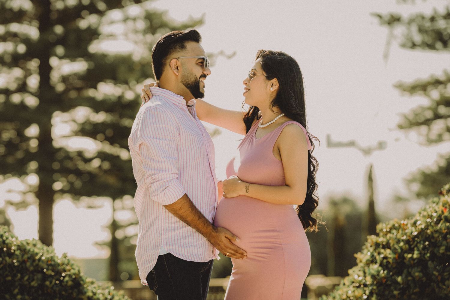 Couple sharing a tender moment outdoors, pregnant woman in pink dress cradling baby bump, partner in pink shirt, trees in background.