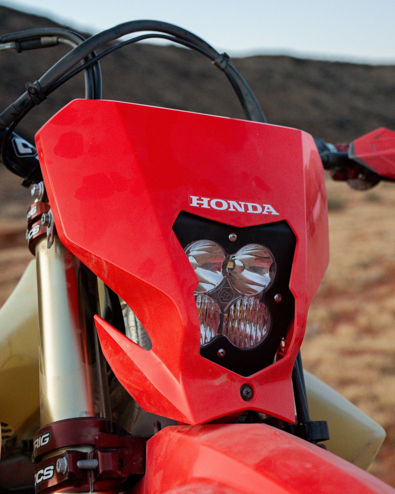 Close-up of a red Honda dirt bike headlight fairing with dual round lights, set against a desert background.