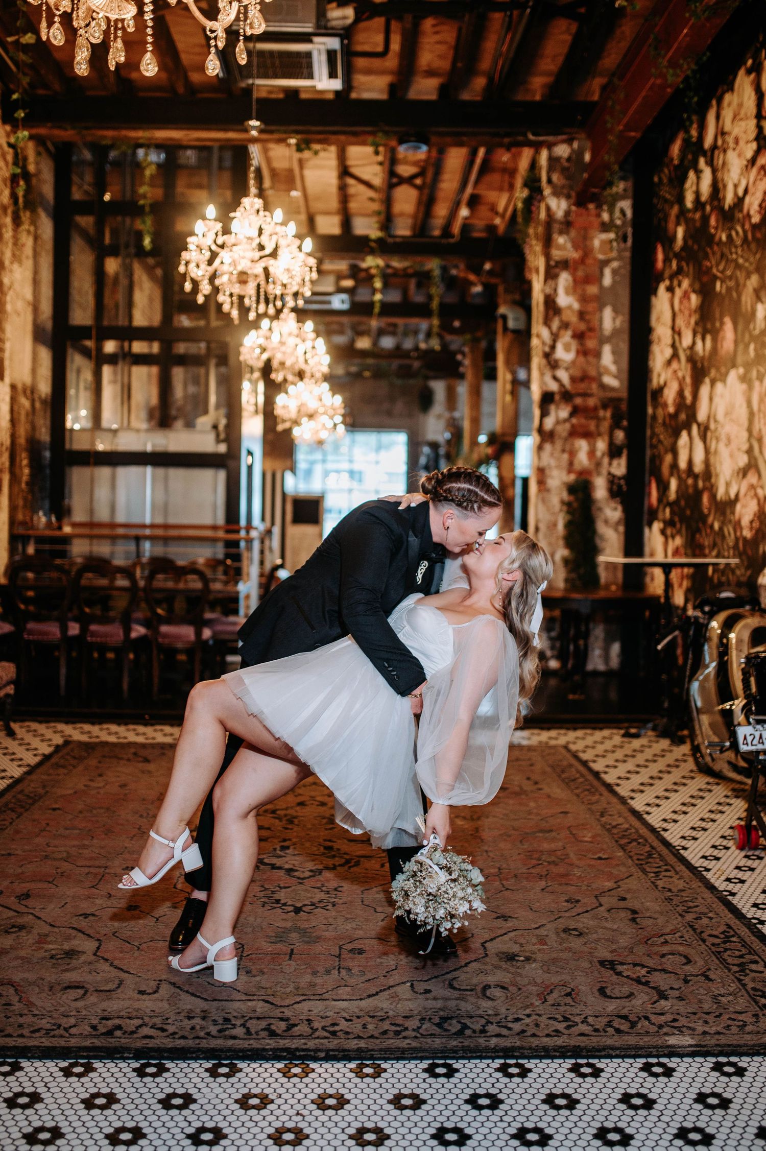 Bride and groom share a romantic dip kiss in an elegant chandelier-lit wedding venue with ornate decor.