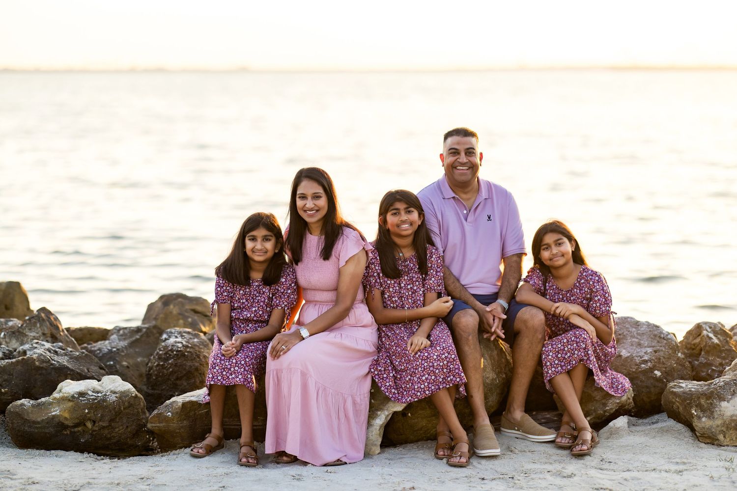Family of five smiling together on rocky beach at sunset, parents with three daughters in matching floral dresses.