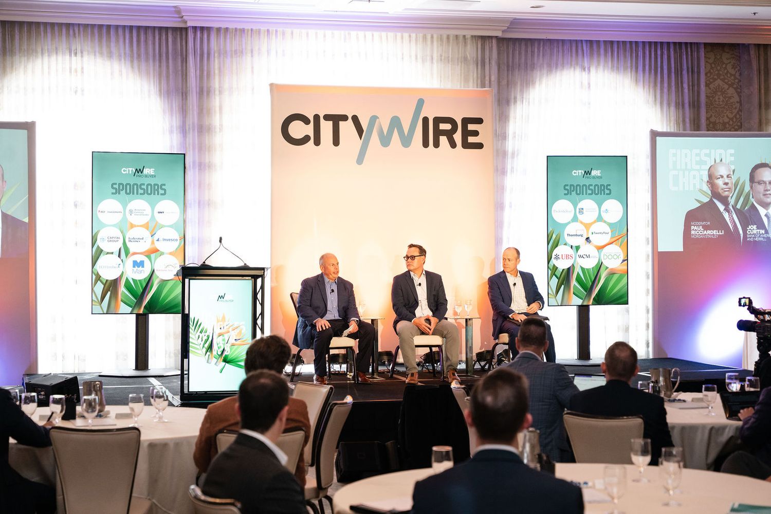 Three panelists speak on stage at a Citywire financial conference in a decorated ballroom with an audience.