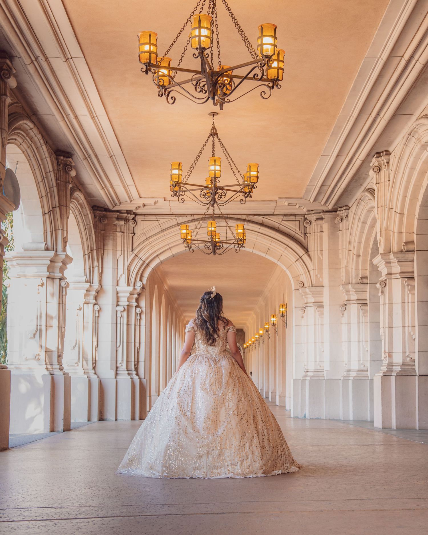 Young woman in a glittering ball gown poses with a book by a grand fountain for her Quinceañera photos.