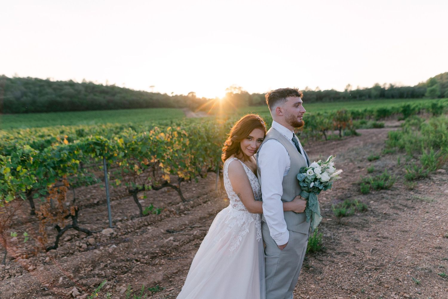 Newlywed couple embraces in sunlit field with wildflowers during golden hour sunset photography session.