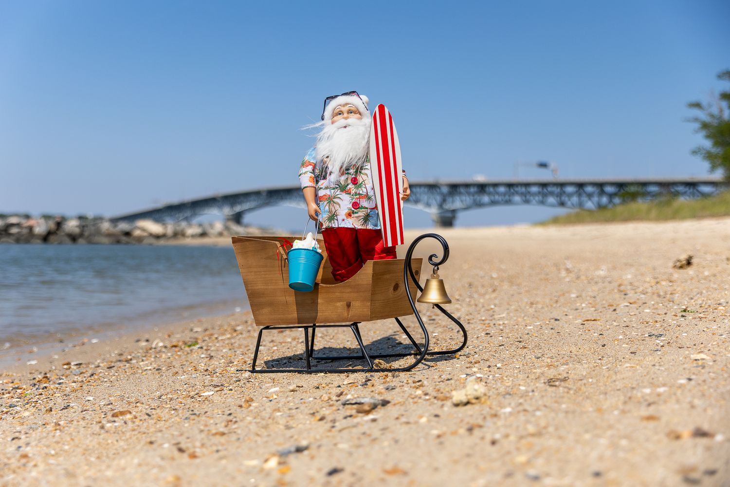 Santa Claus figurine sitting in a sleigh on a sunny beach with a bridge and river in the background.
