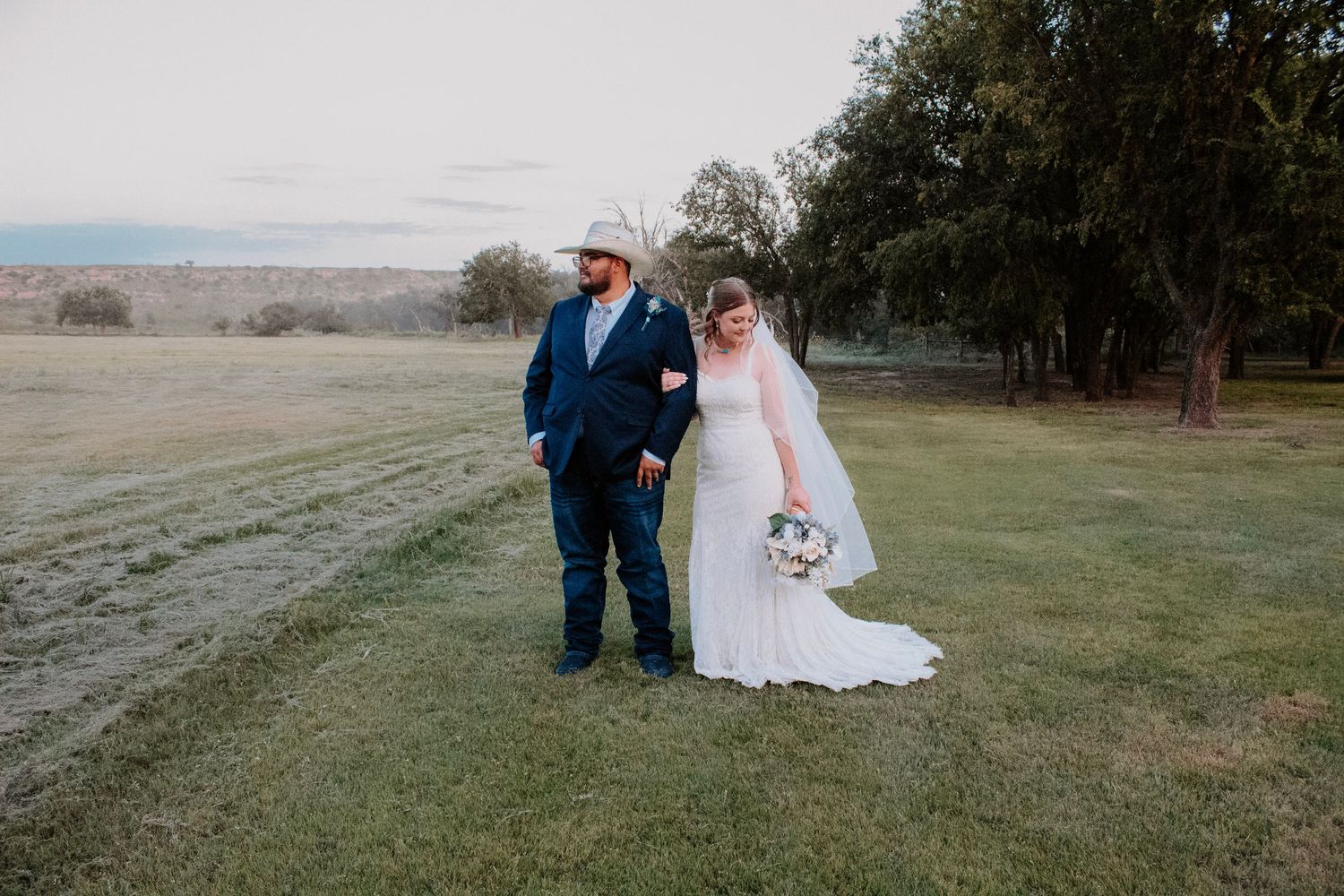 A bride in a white gown and groom in a navy suit hold hands in a lush green field at dusk.