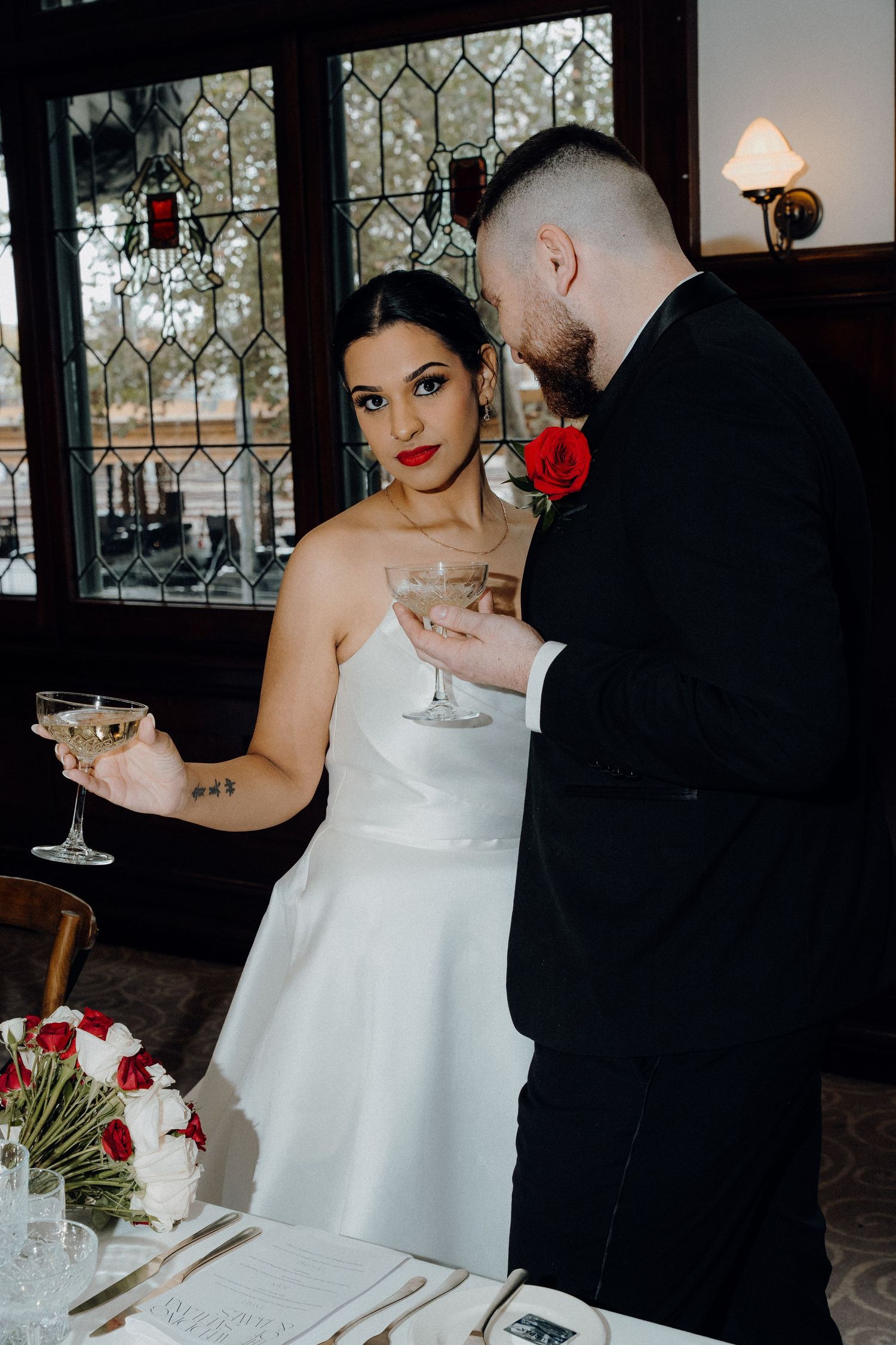 Bride and groom toasting with champagne glasses at wedding reception, bride in white gown with red lips, groom in black suit.
