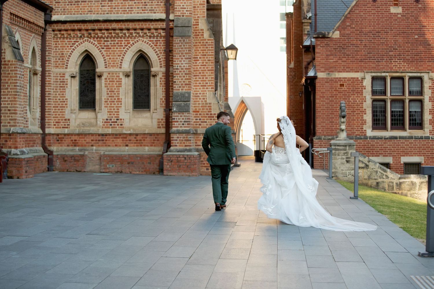 Bride and groom walking hand in hand past a historic red brick church on their wedding day.