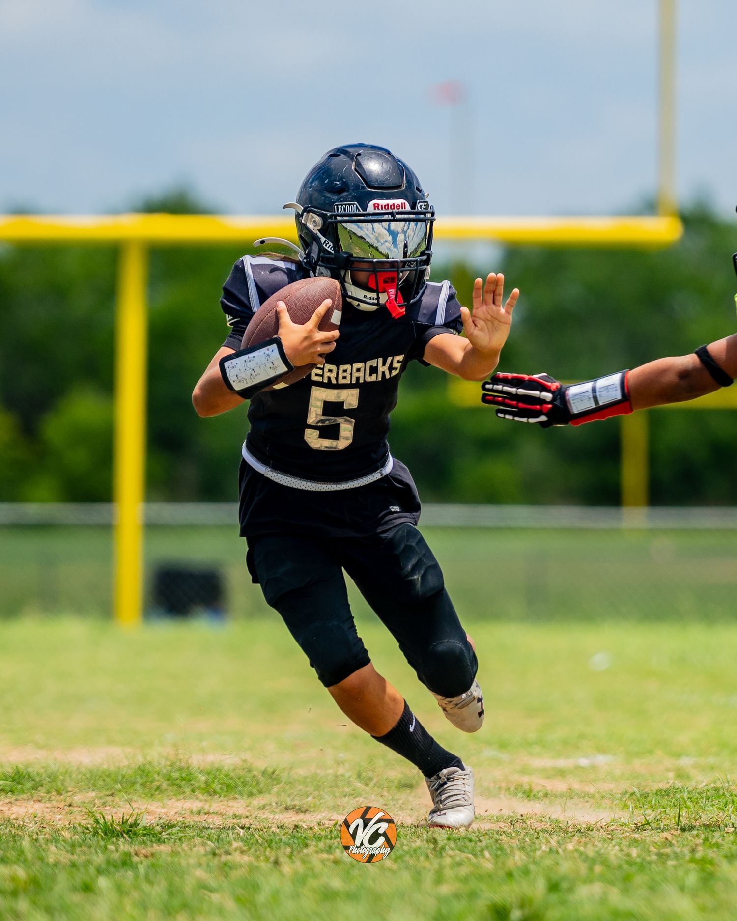 Young football player #5 in black Razorbacks uniform runs with ball during youth league game on green field.