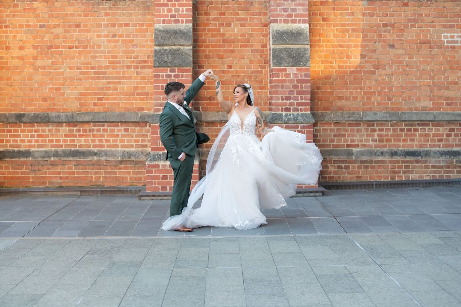 Bride in flowing white gown twirls with groom in green suit against rustic brick wall backdrop.
