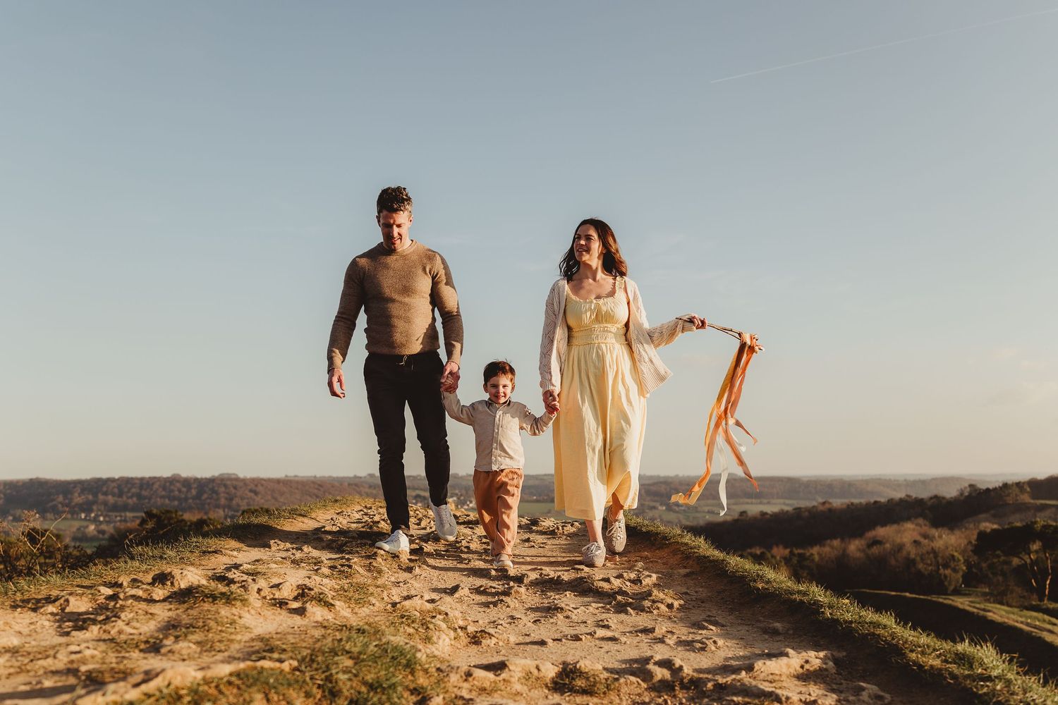 Family of three walking on a rocky hilltop at golden hour, parents holding child's hands against a clear blue sky.