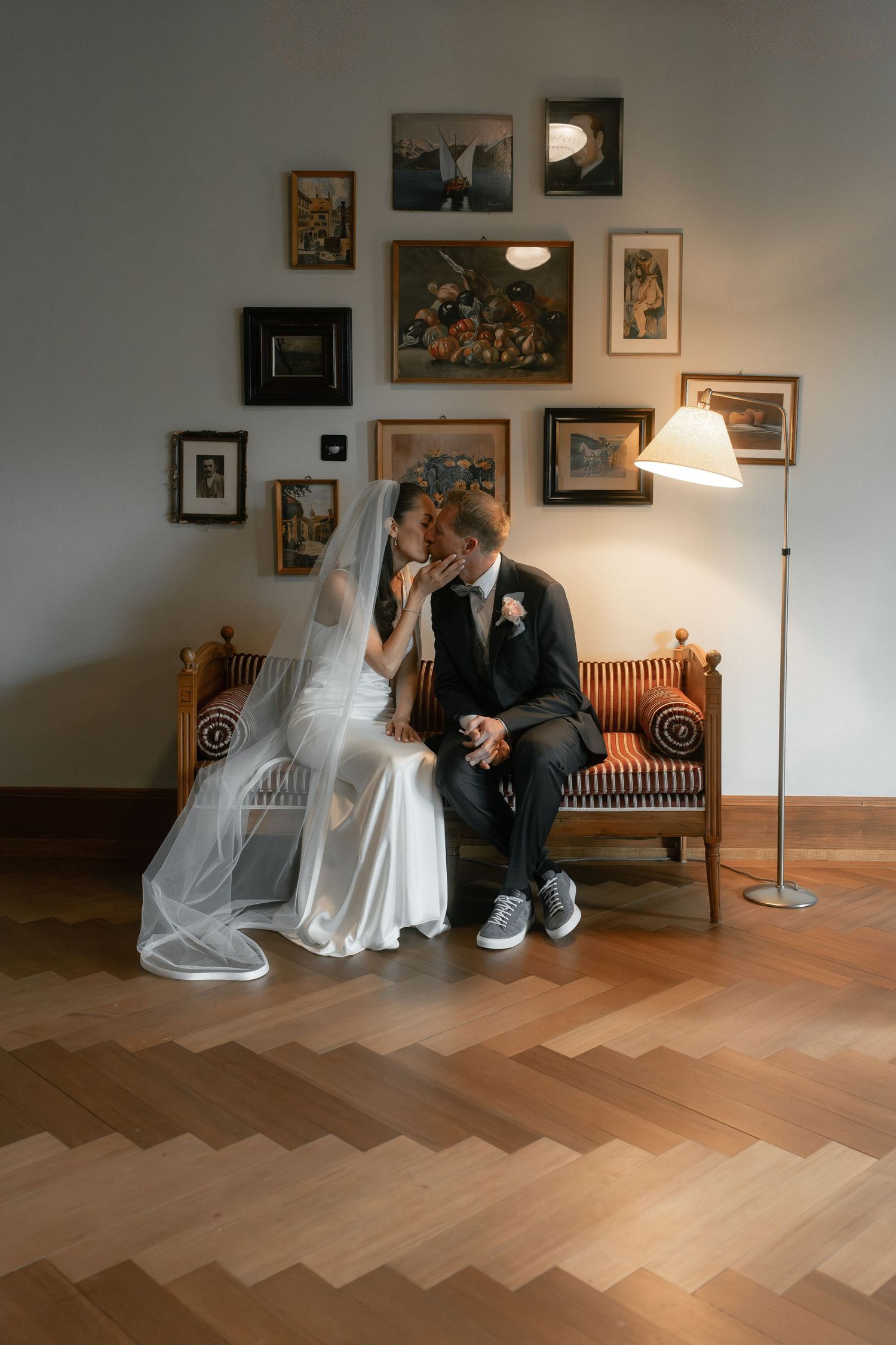 Bride and groom share a kiss on an ornate bench surrounded by gallery wall art and warm lamp light on herringbone floor.