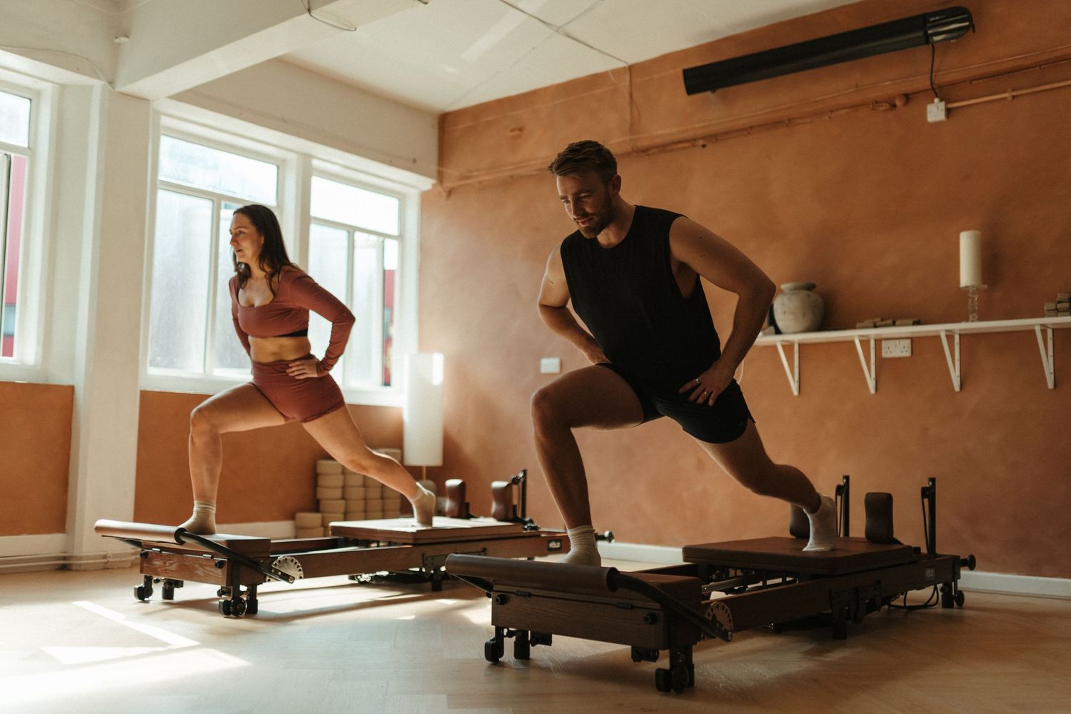 Man and woman performing lunges on Pilates reformers in a bright modern fitness studio.