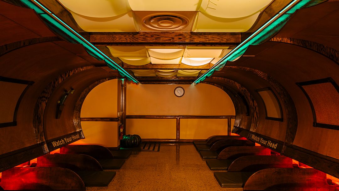 Art Deco subway station interior with arched ceiling, neon lighting, and rows of benches in warm amber tones.