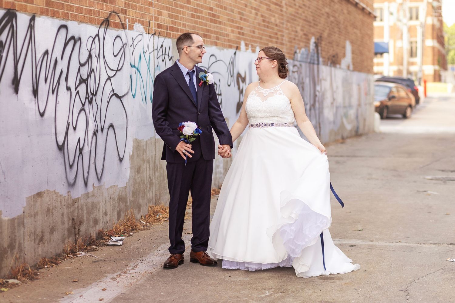 Bride and groom holding hands near graffiti wall on wedding day in urban outdoor setting.