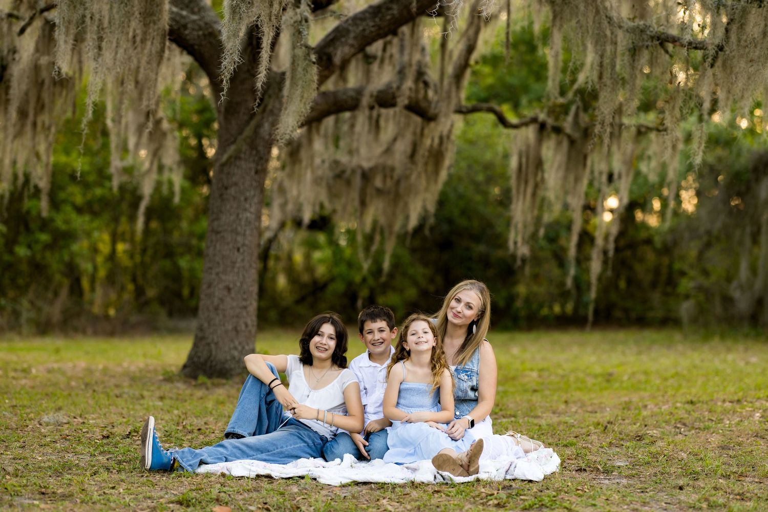 Family of four smiling together on a blanket under a moss-draped oak tree in a lush green park.
