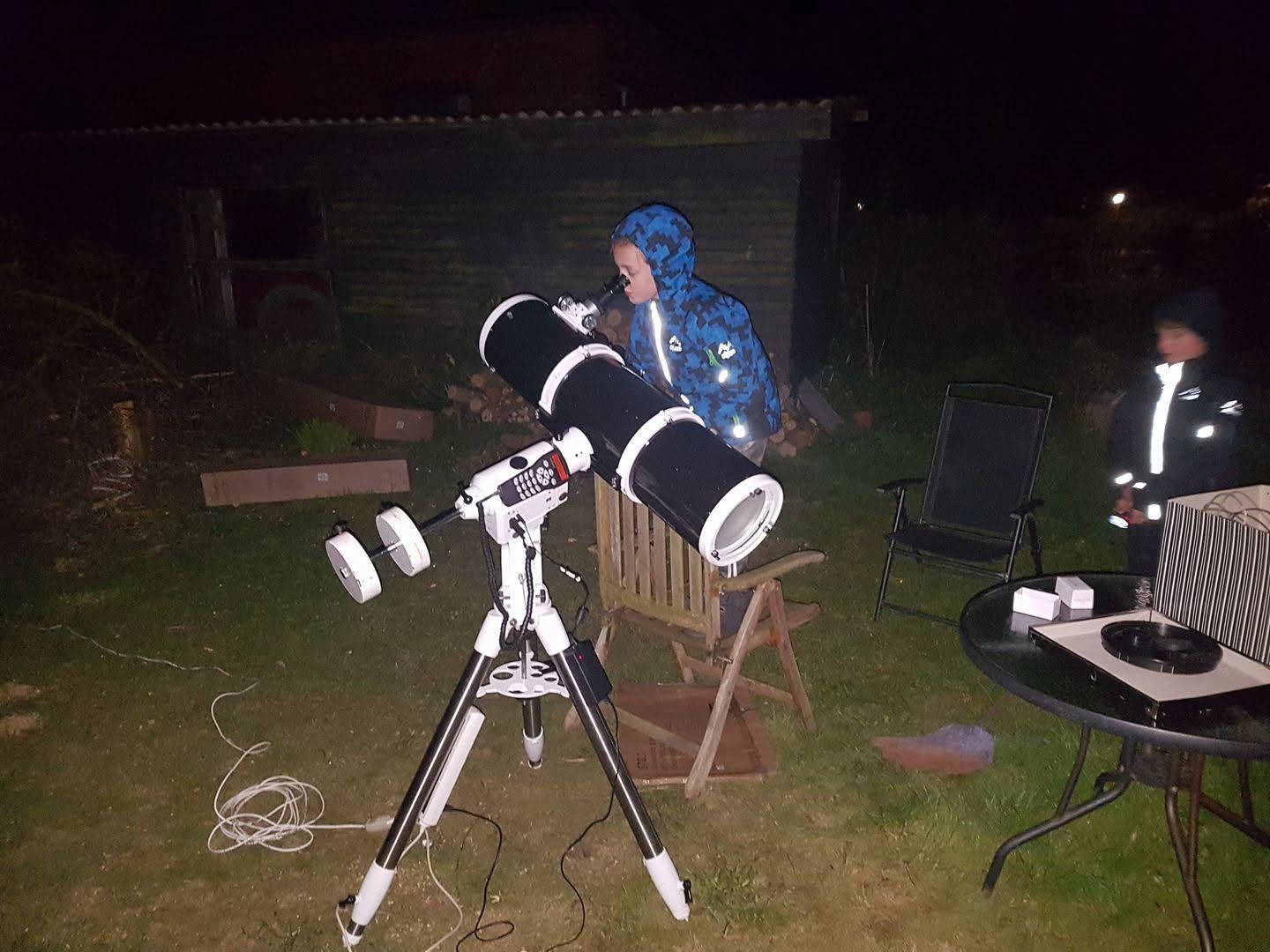 Person in blue jacket using a large telescope on a tripod in a dark backyard at night for stargazing.