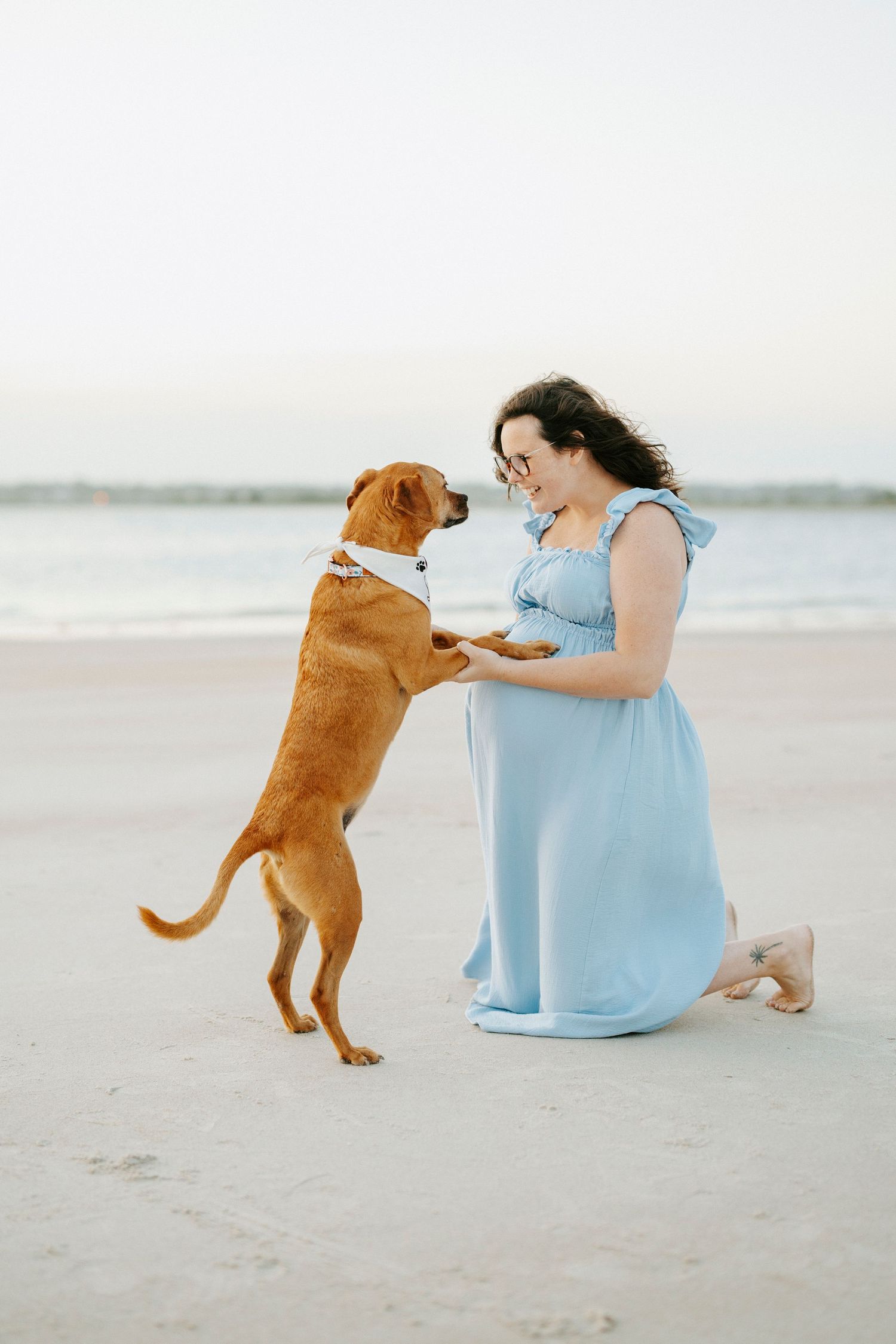 Woman in blue dress kneeling on beach, holding paws with brown dog wearing white bow tie at sunset.