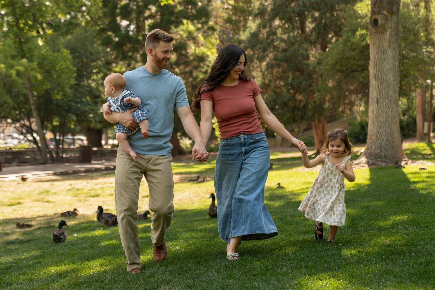 Family of four walking in a sunny park, parents holding hands with toddler girl while dad carries baby.