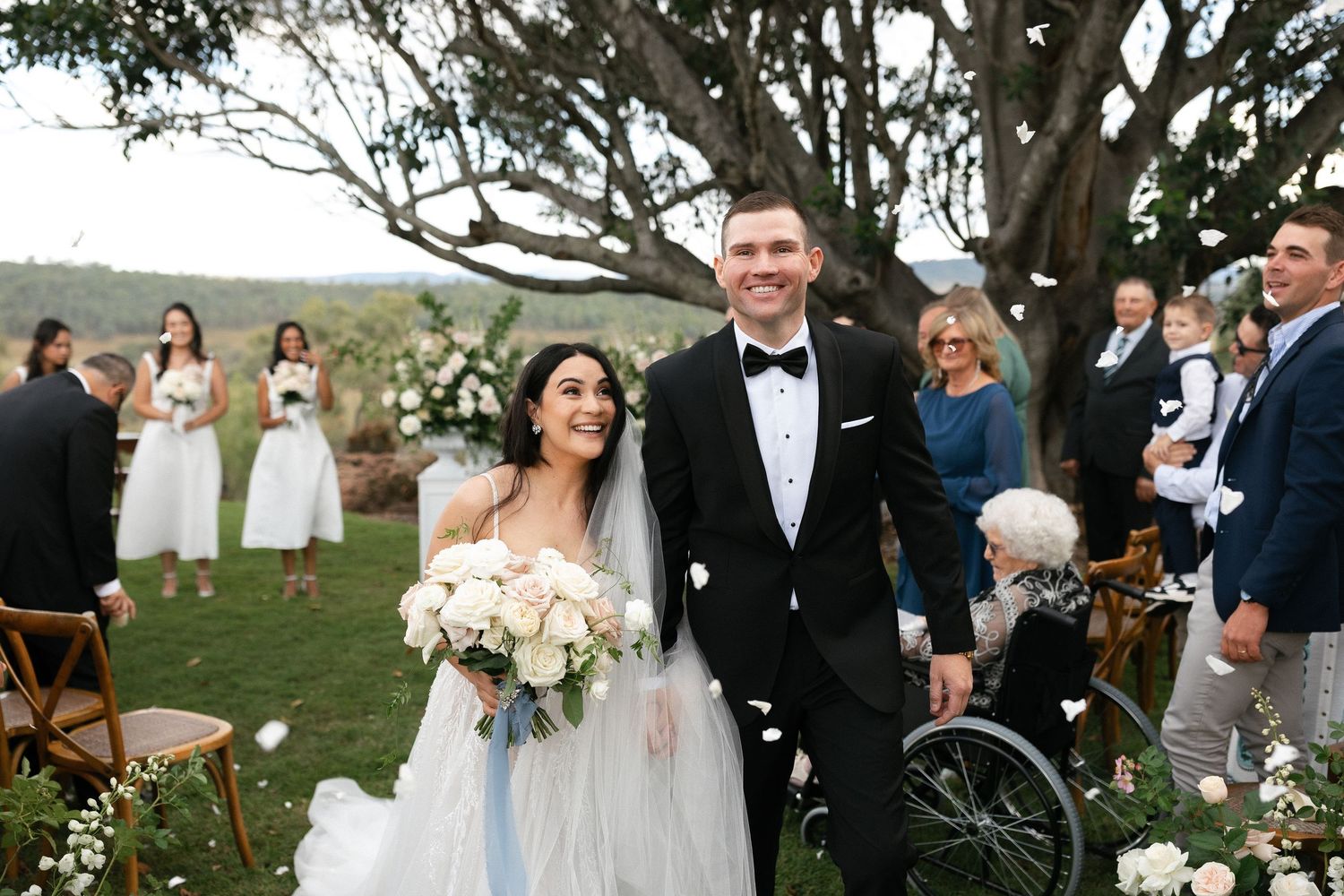 Bride and groom walk down the aisle smiling, surrounded by guests outdoors under a large tree with white floral decor.