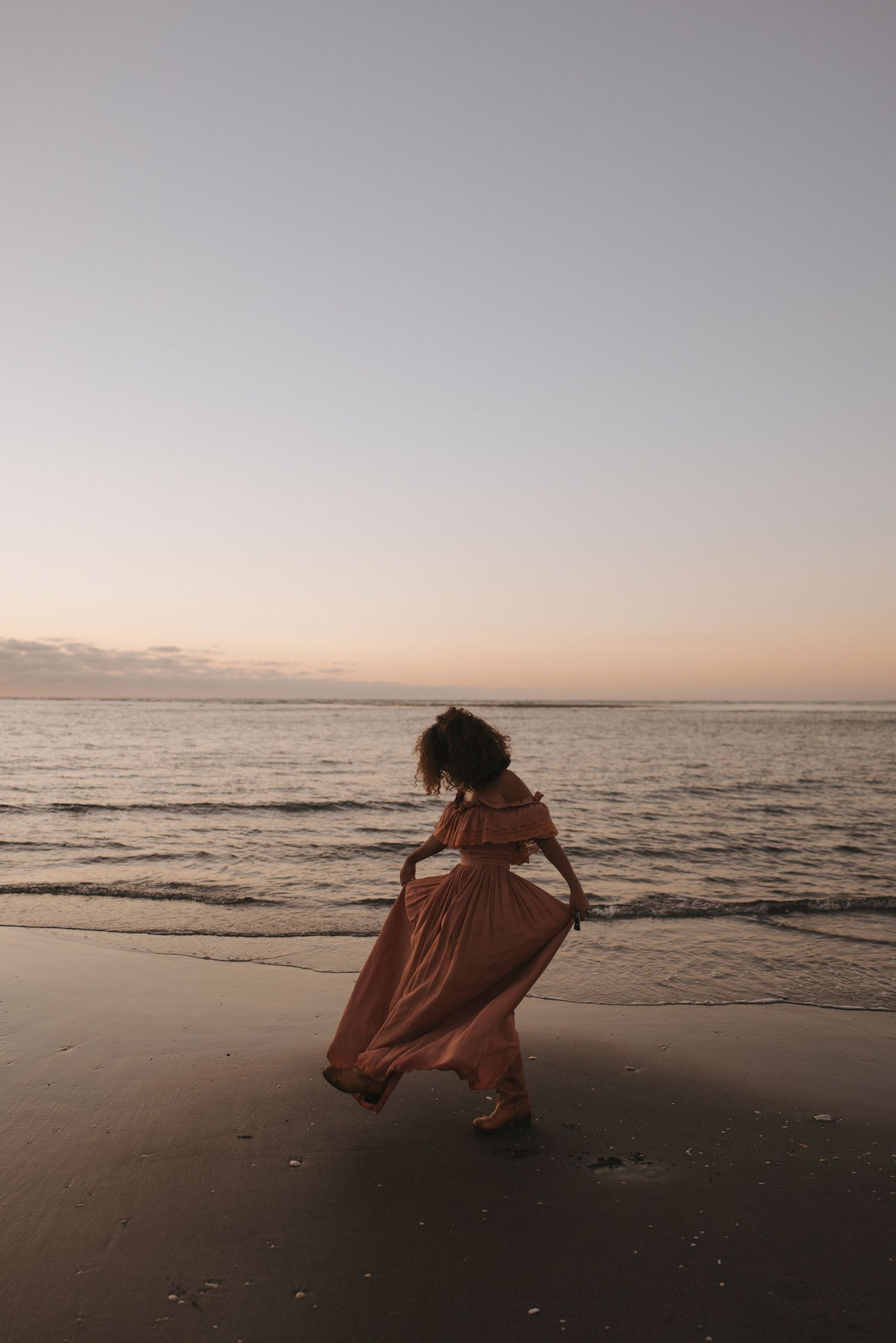 A woman in white stands on a sandy beach with arms raised, under a dramatic cloudy sky near the ocean.
