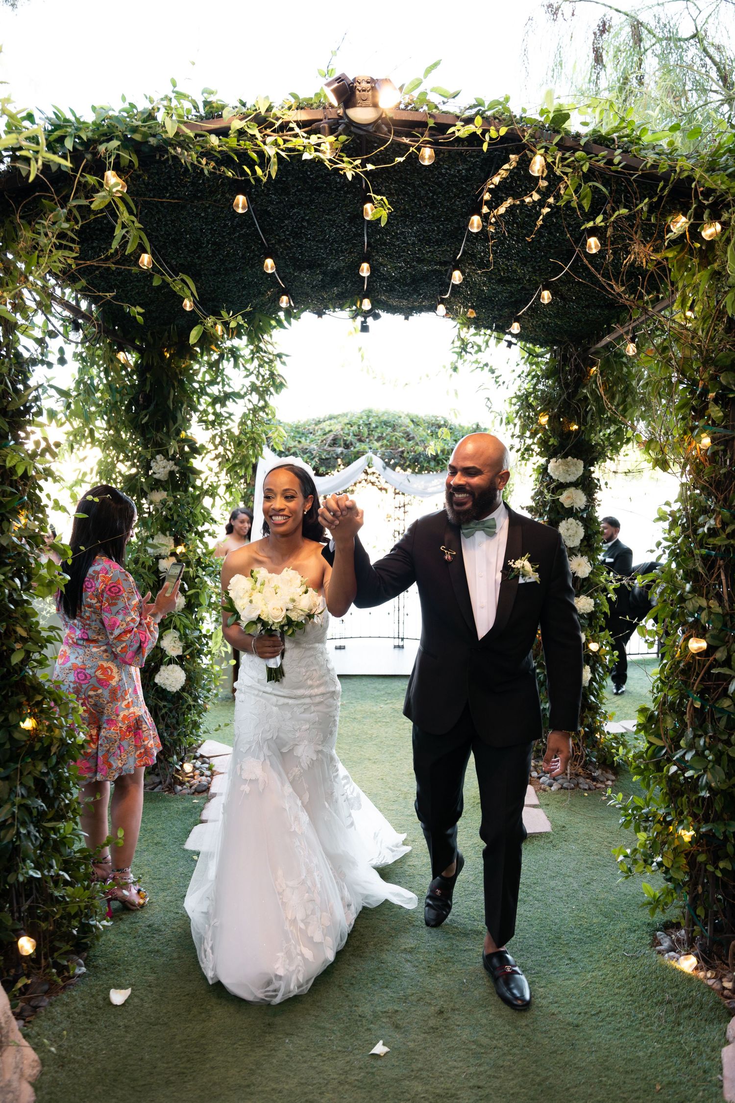 Joyful newlywed couple walks down the aisle after outdoor wedding ceremony, bride holding white bouquet.