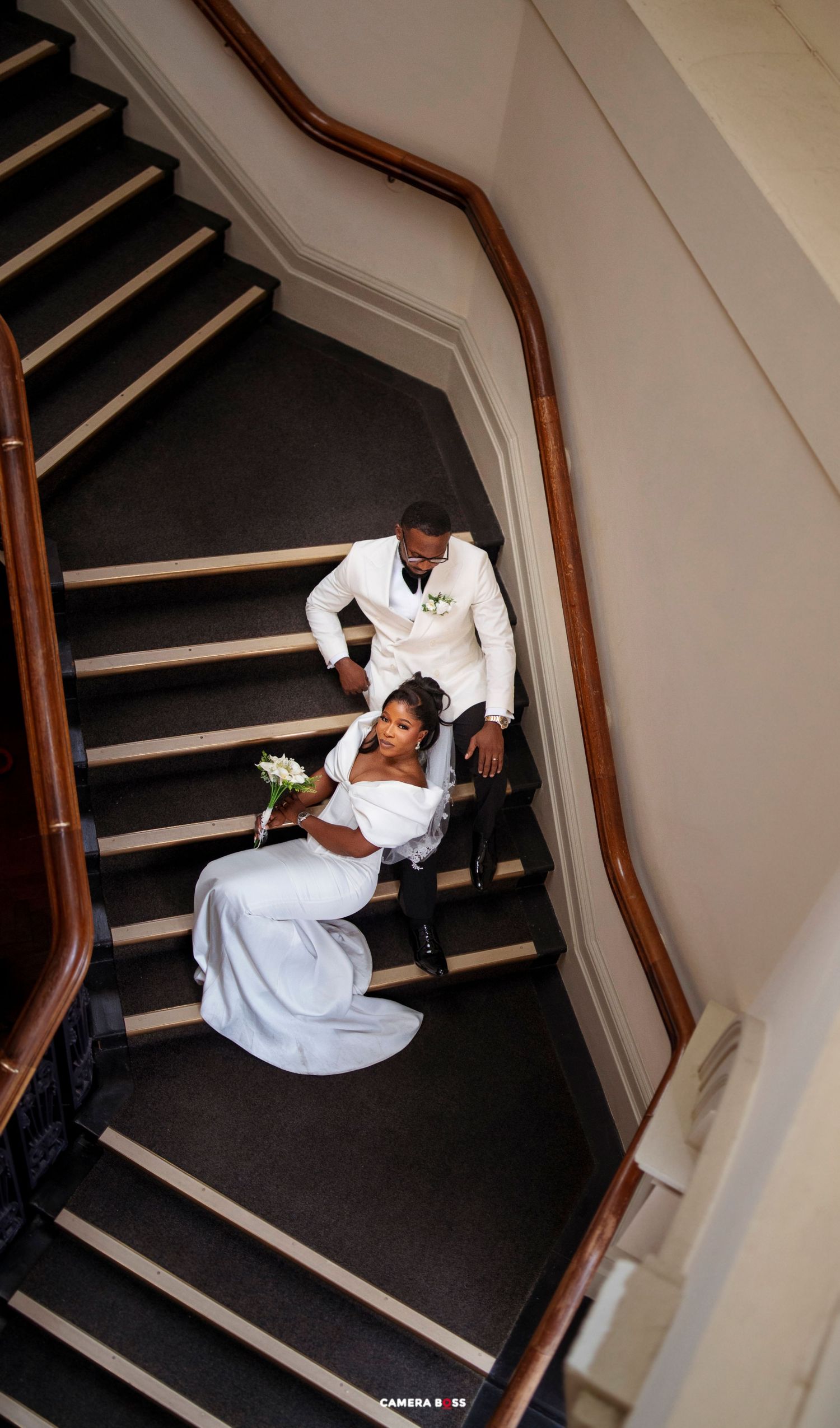 Aerial view of a bride in white gown and groom in white suit posing elegantly on a grand staircase.