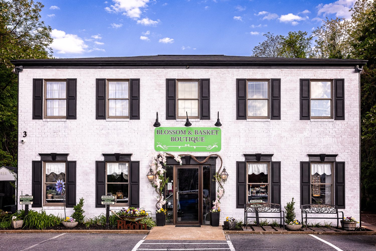 White two-story boutique building with black shutters, green awning sign, and charming storefront decor surrounded by trees.
