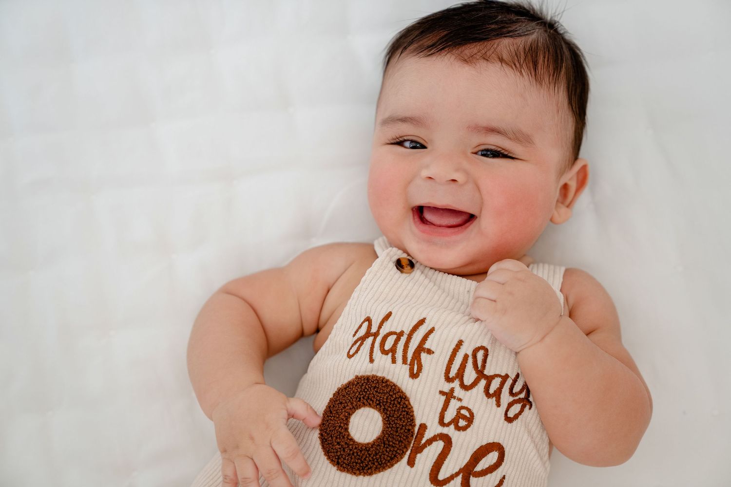 Happy baby wearing white onesie with brown text design lies on white blanket while smiling at camera.